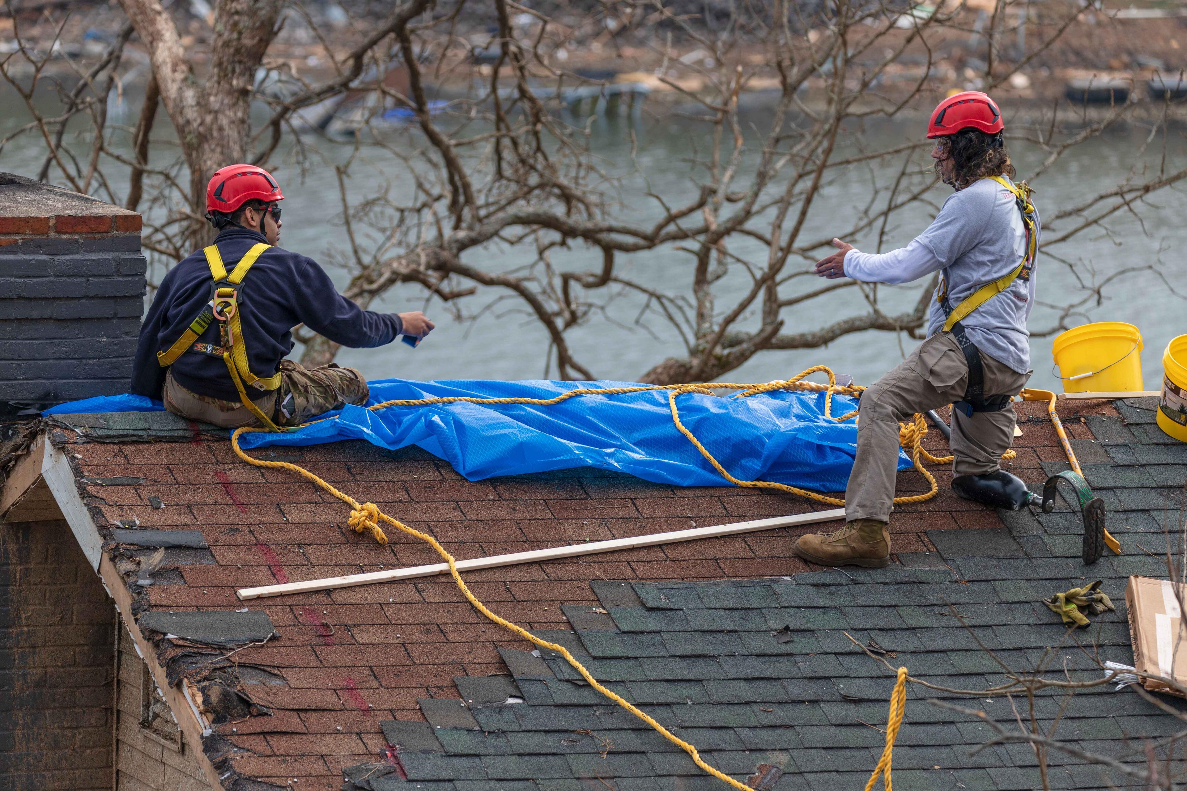 Two Greyshirt volunteers sit on the roof of a home. The roof's shingles have fallen off due to a storm. In the background there are dead trees and a small lake.